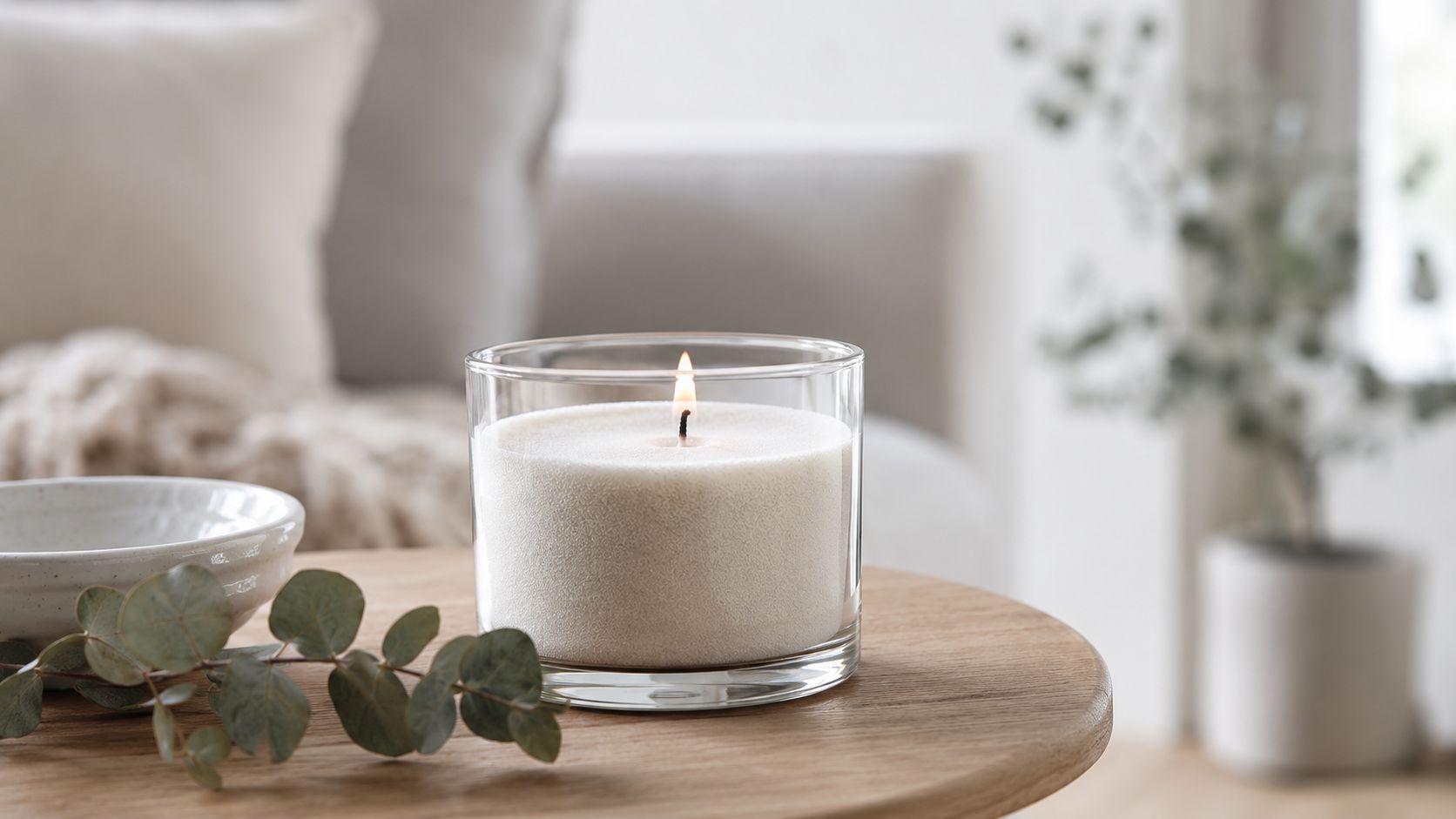 A lit white candle in a glass jar sitting on a wooden table with eucalyptus sprigs.